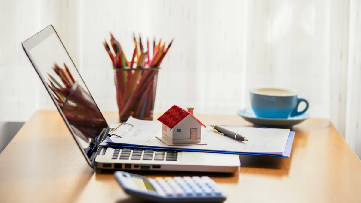 Desk with a laptop, calculator, small model of a home, pencil cup, and a cup of coffe