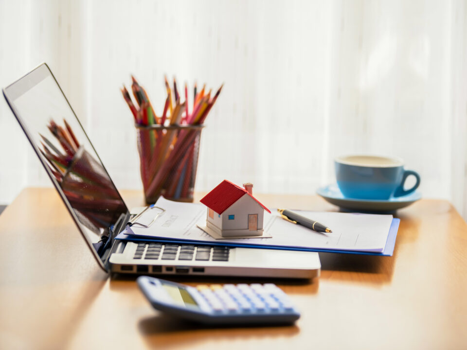 Desk with a laptop, calculator, small model of a home, pencil cup, and a cup of coffe